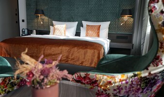 Una camera d'albergo con un letto grande, due lampade sui comodini, una poltrona in tessuto verde e un'esposizione floreale.