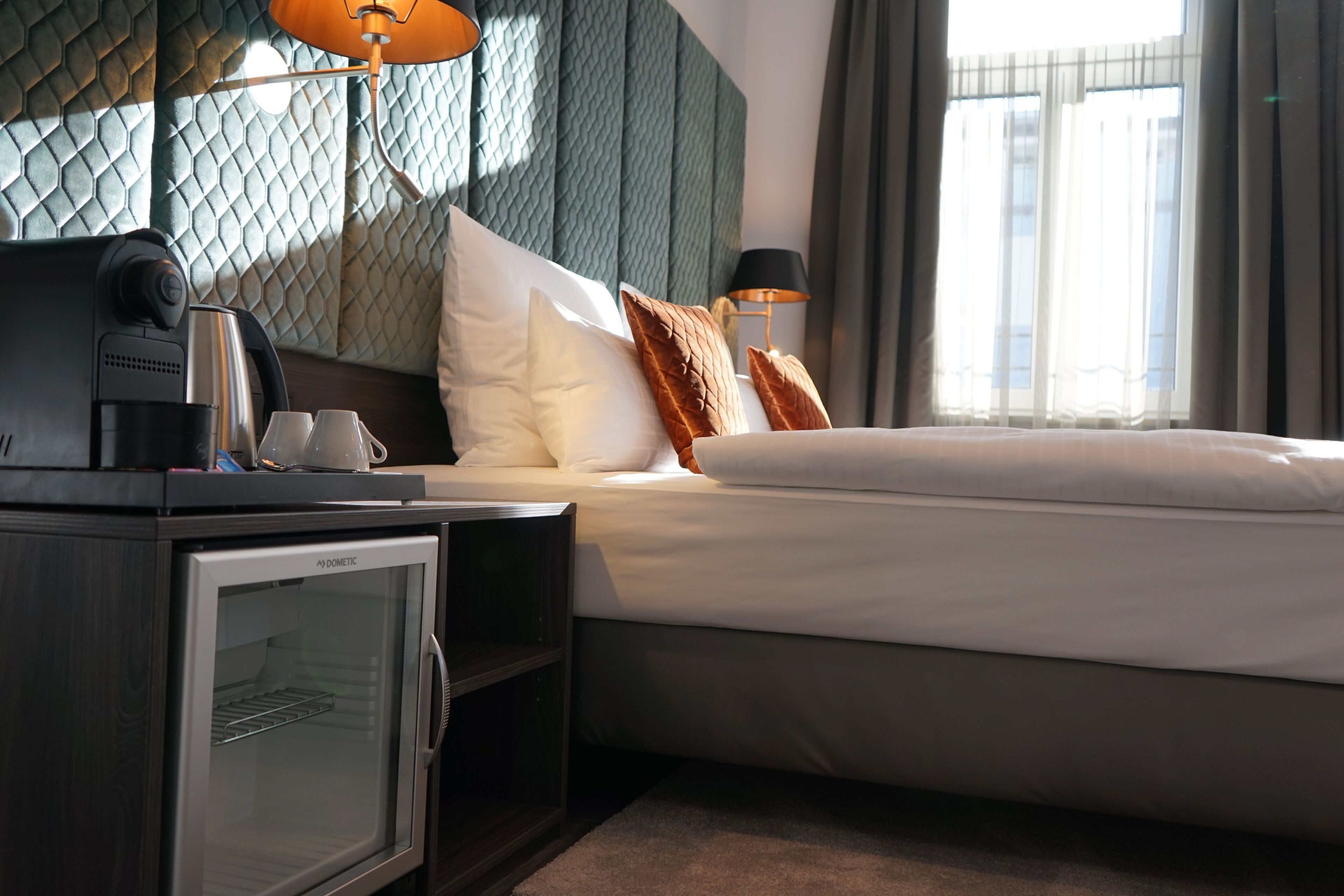 Ein Hotelzimmer mit Bett, Minibar, Lampe und Blick auf die Stadt durch das Fenster.