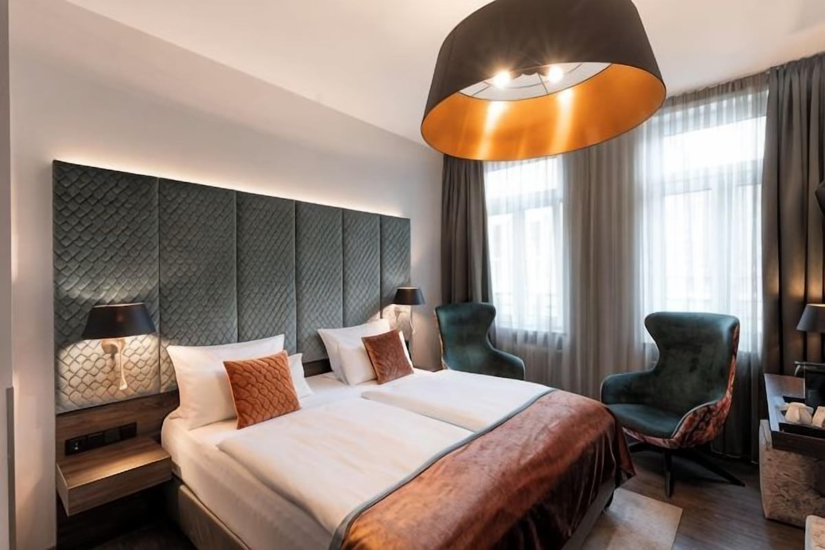 A hotel room with a large bed, two bedside tables, a green armchair, a desk with a lamp, and a large window with curtains.