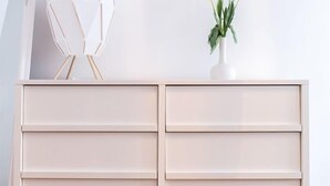 Un tocador blanco minimalista con tres cajones, una escultura blanca de formas geométricas y un florero con flores blancas encima.
