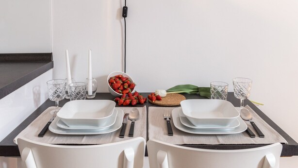 Una mesa de comedor dispuesta con platos blancos, cubiertos y vasos, con un cuenco de fresas y un bulbo de ajo sobre la mesa.