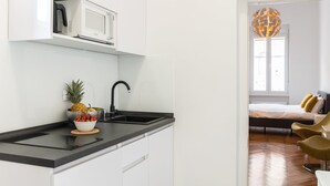 Una cocina moderna con armarios blancos, encimera negra y microondas. Hay un cuenco de fruta en la encimera y un lavabo con grifo negro. El piso es de madera y se ve una cama con almohadas amarillas al fondo.