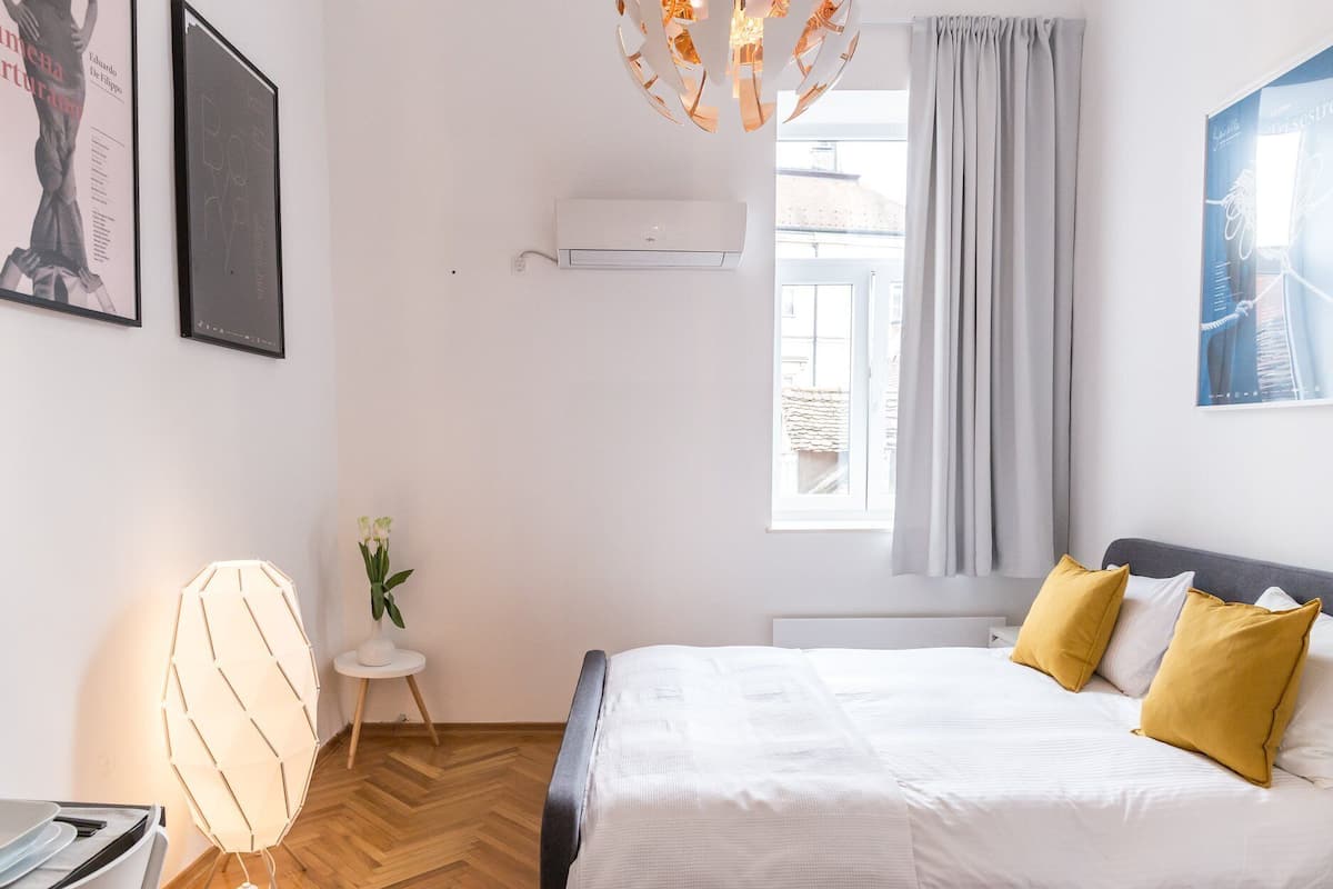 A bedroom with a bed, a small table with a plant, a wall-mounted air conditioner, and a window with curtains.