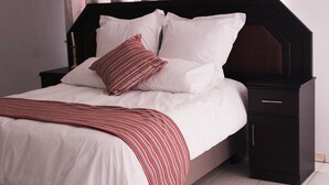 A neatly made bed with white linens and a striped throw blanket, a dark wooden headboard, and a small bedside table with a drawer. - Flamboyant Place (Harare)