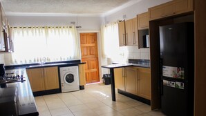 A kitchen with wooden cabinets, a black refrigerator, a washing machine, and a window with curtains. - Flamboyant Place (Harare)