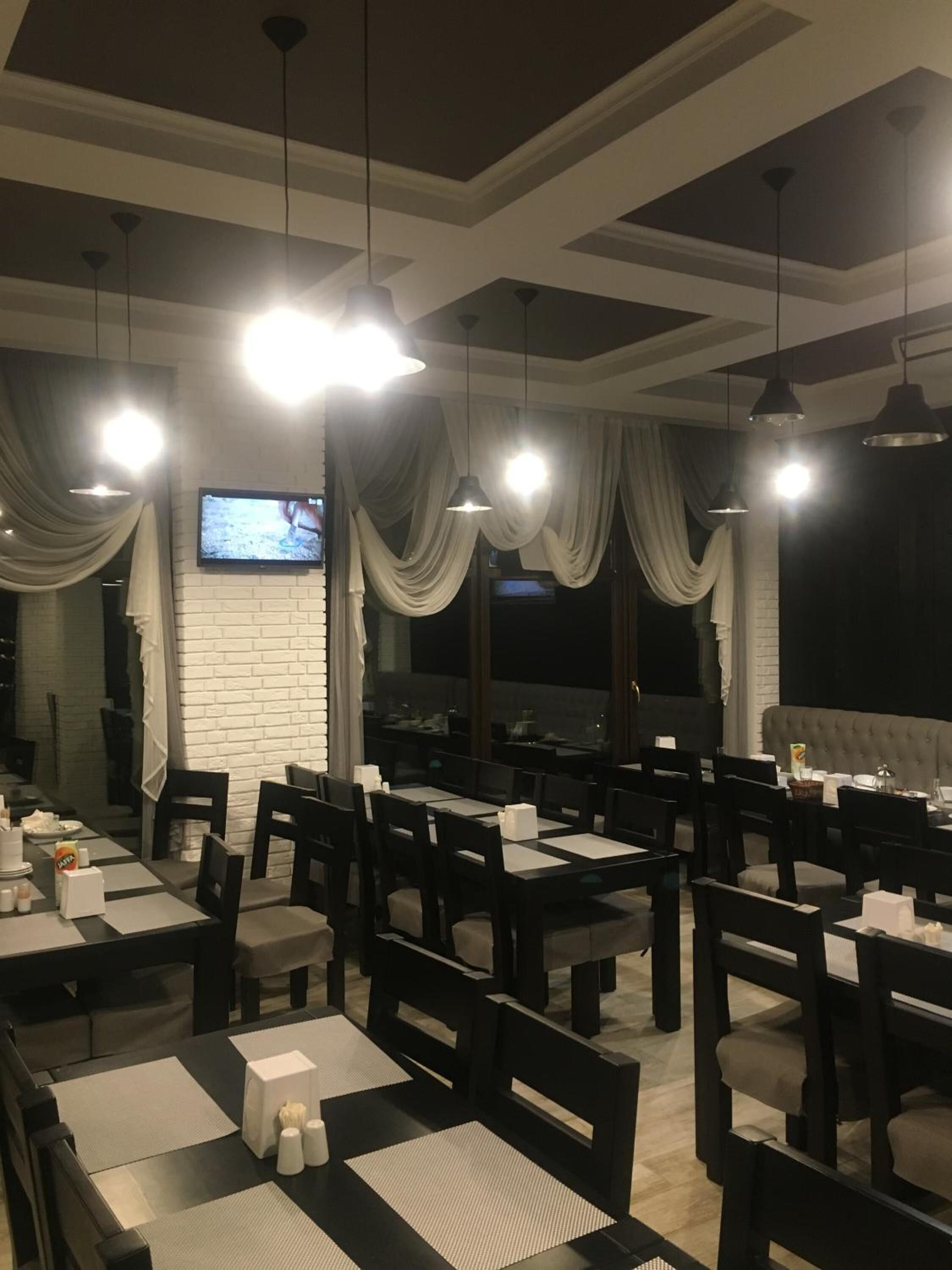 A restaurant with multiple tables and chairs, a television screen, and draped curtains.