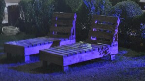 A wooden bench with a backrest and armrests, placed on a grassy area with bushes in the background.