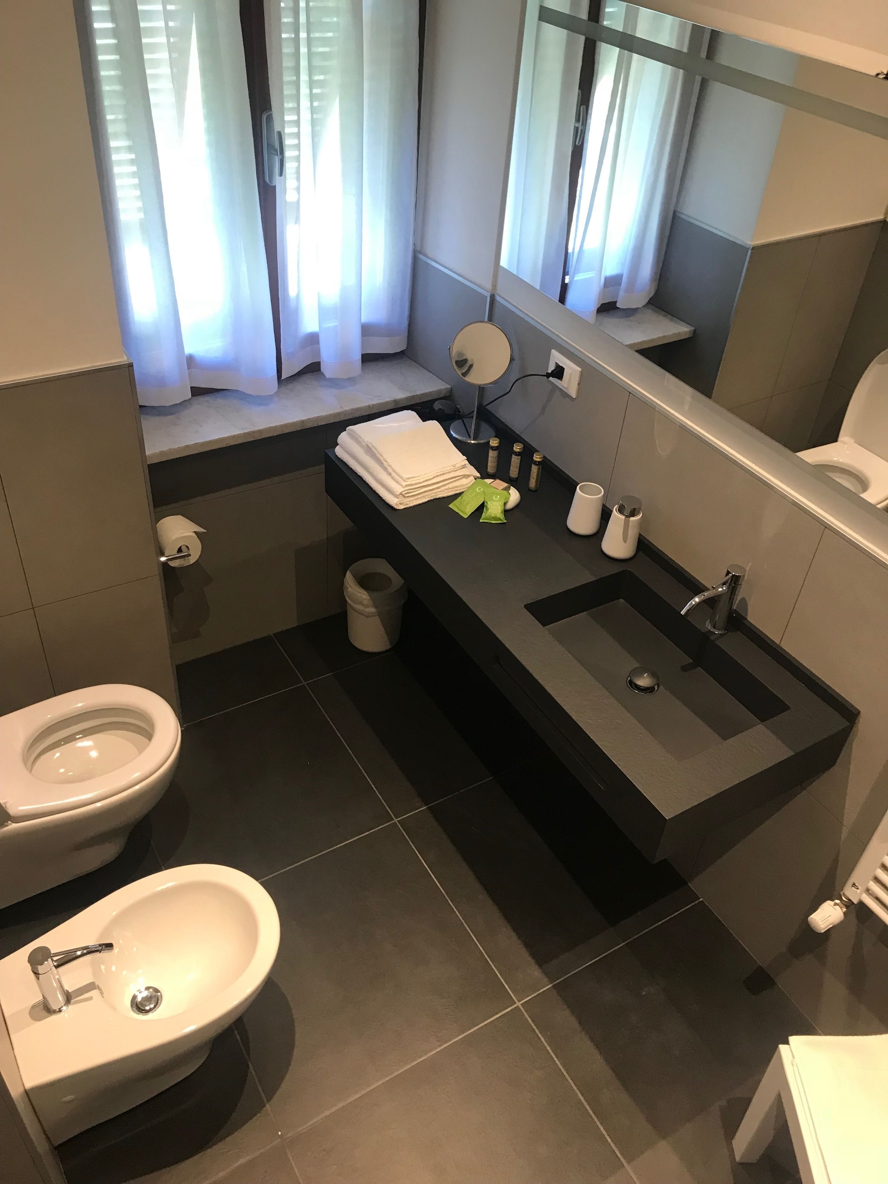 A modern bathroom with a toilet, bidet, and sink. There is a window with curtains, a mirror, and toiletries on the sink counter.