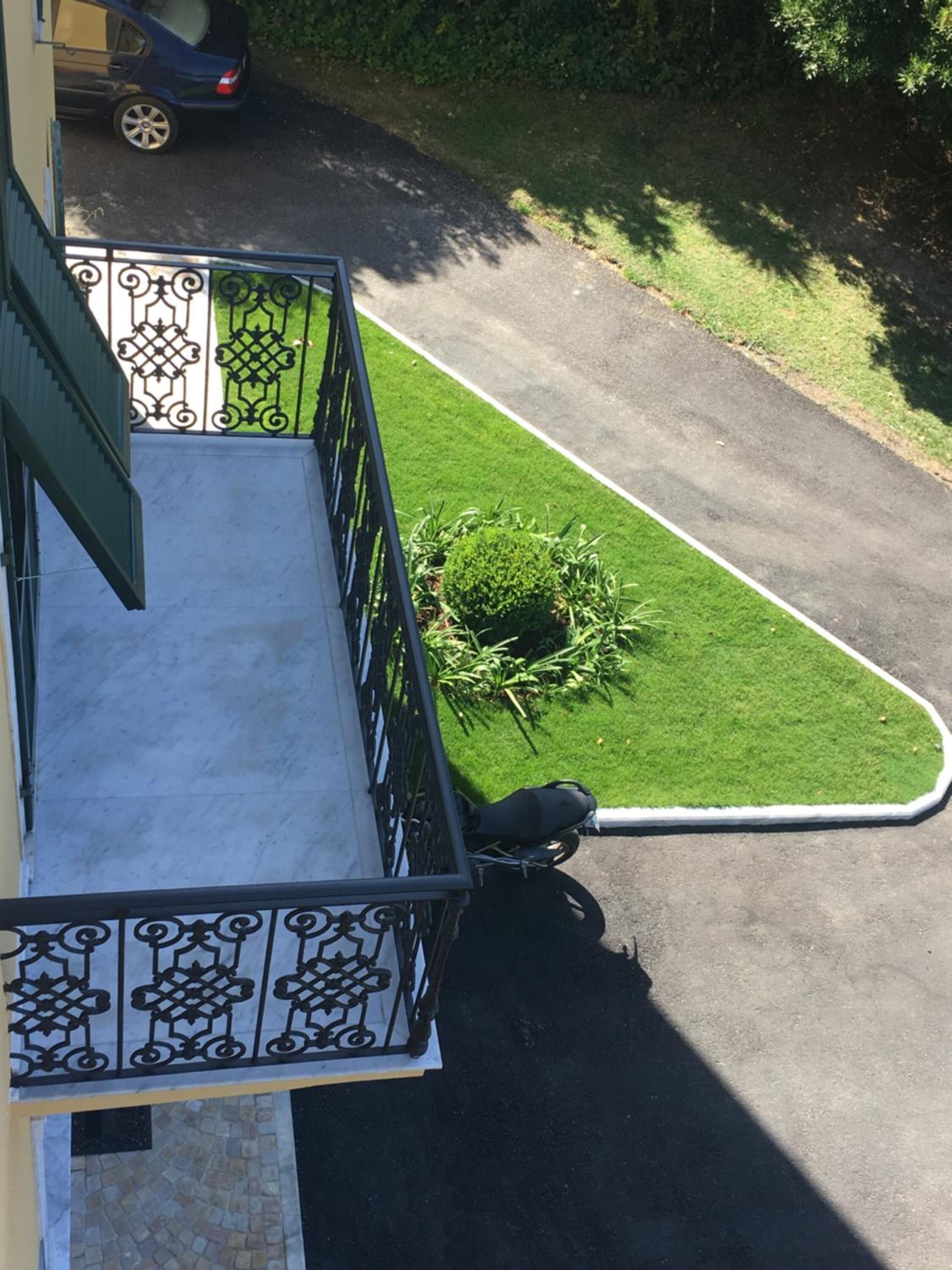 A balcony with a metal railing, a small garden area with a circular hedge, and a parked car in the background.