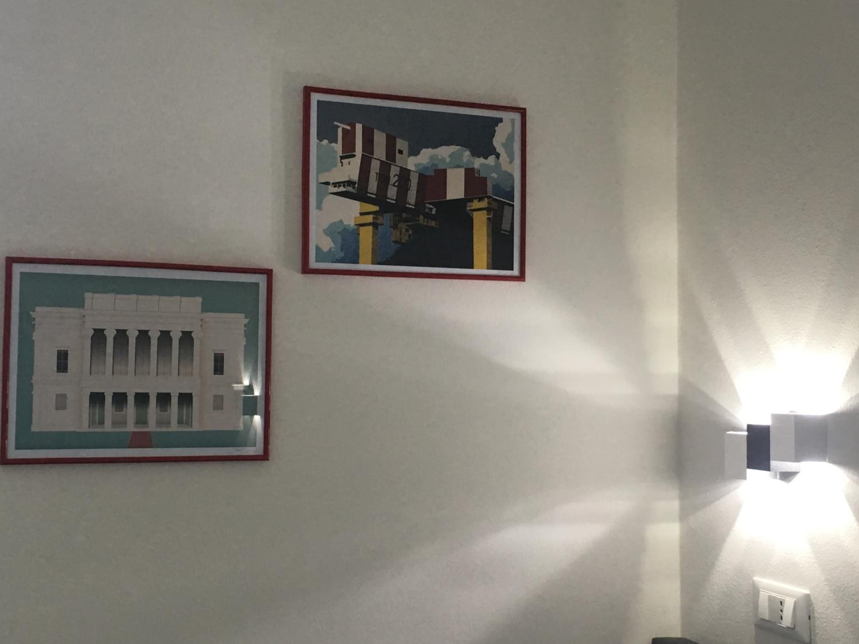 Twee ingekaderde foto's aan een muur, één toont een gebouw met kolommen en de ander een constructie met schoorstenen.