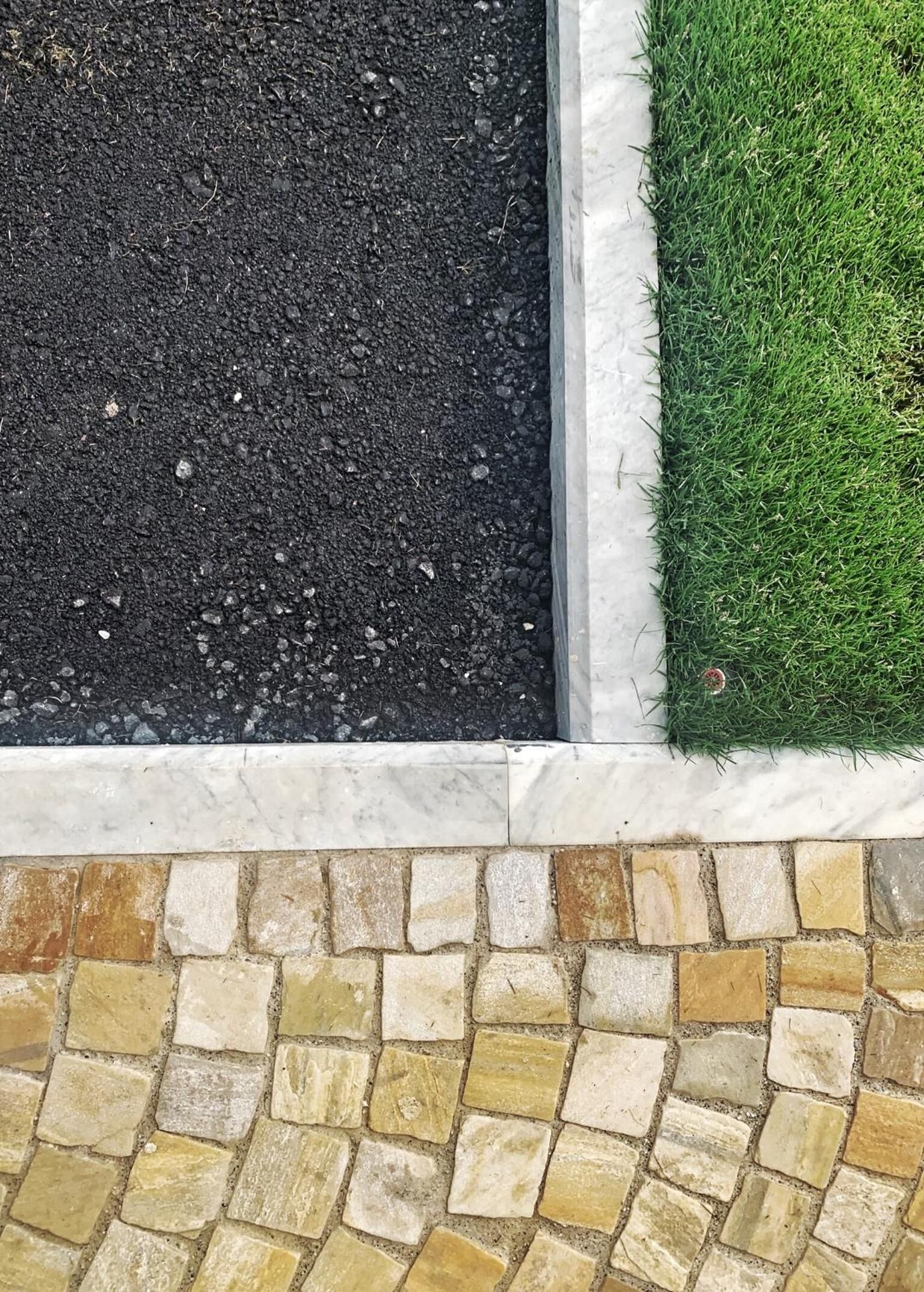 Ein Blumenbeet mit dunkler Erde und grünem Gras, begrenzt durch eine weiße Steinmauer und eine Mosaikfliesen-Unterlage.