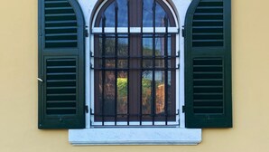 A window with green shutters and a white arch above it, set in a yellow wall with a blue baseboard. - B&B Mikeme (Carrara)