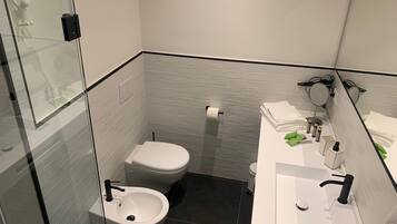 Un baño moderno con mampara de vidrio, inodoro, bidé, lavamanos con espejo y toallas ordenadas.