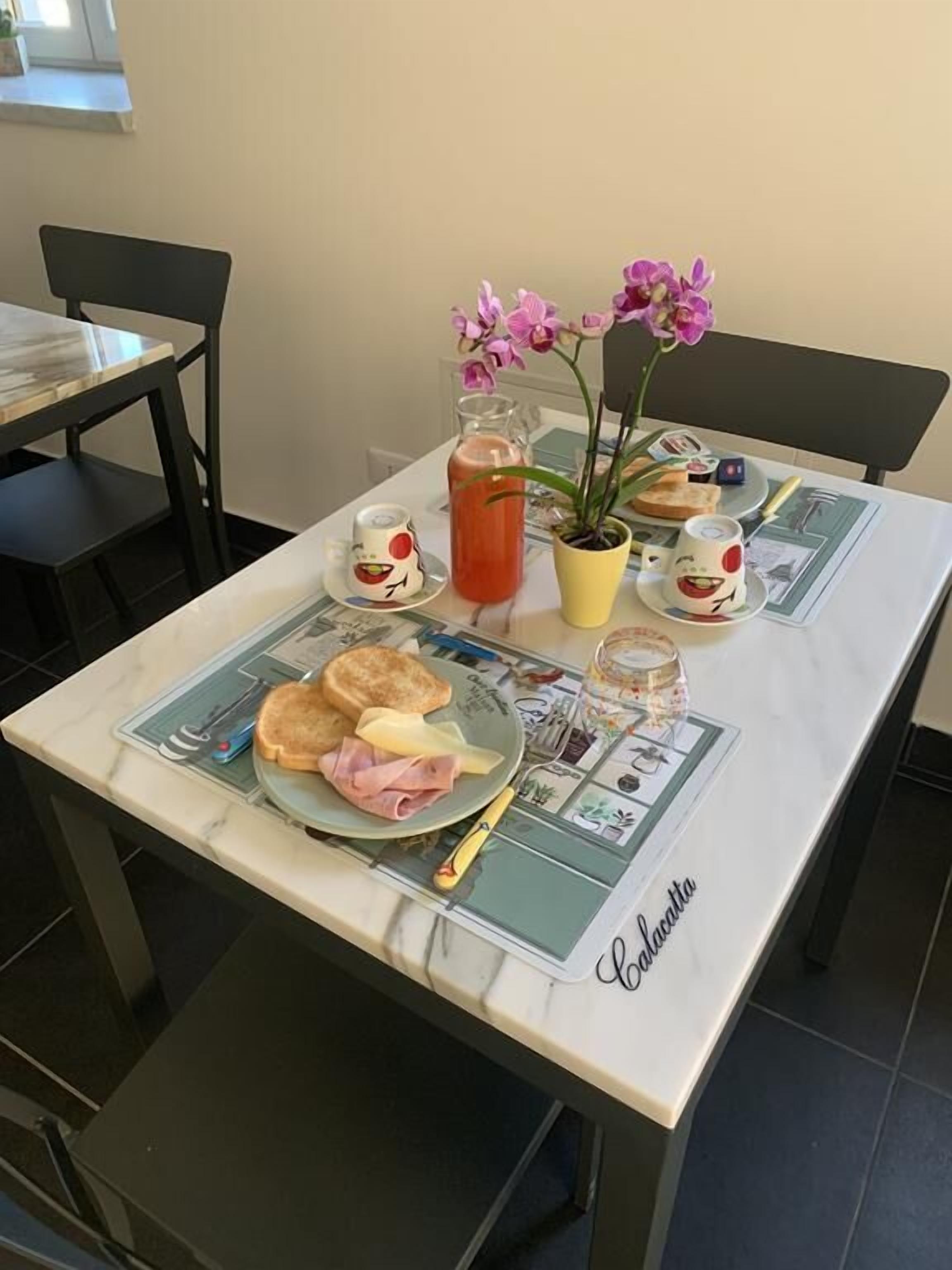 A dining table with a breakfast setting including toast, ham, and eggs, a pitcher of juice, and a potted orchid.