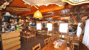 Ein rustikales Holzinterieur mit mehreren Esstischen und Stühlen, einer Küchenzeile mit hängendem Kochgeschirr sowie einer Decke mit freiliegenden Balken und einer gewebten Leuchte.