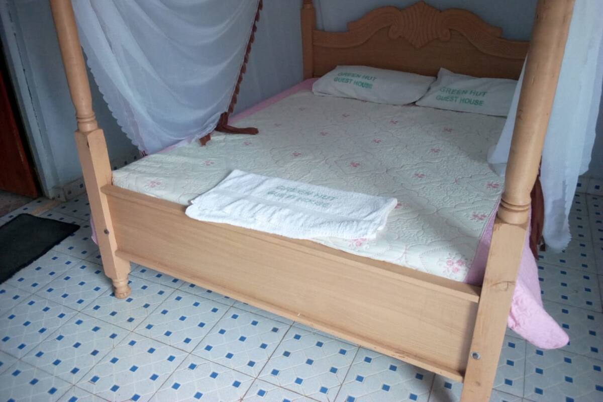 Nelipylväinen sänky, jossa on valkoinen peitto ja tyynyissä lukee teksti: ”GREEN HUT GUEST HOUSE”.