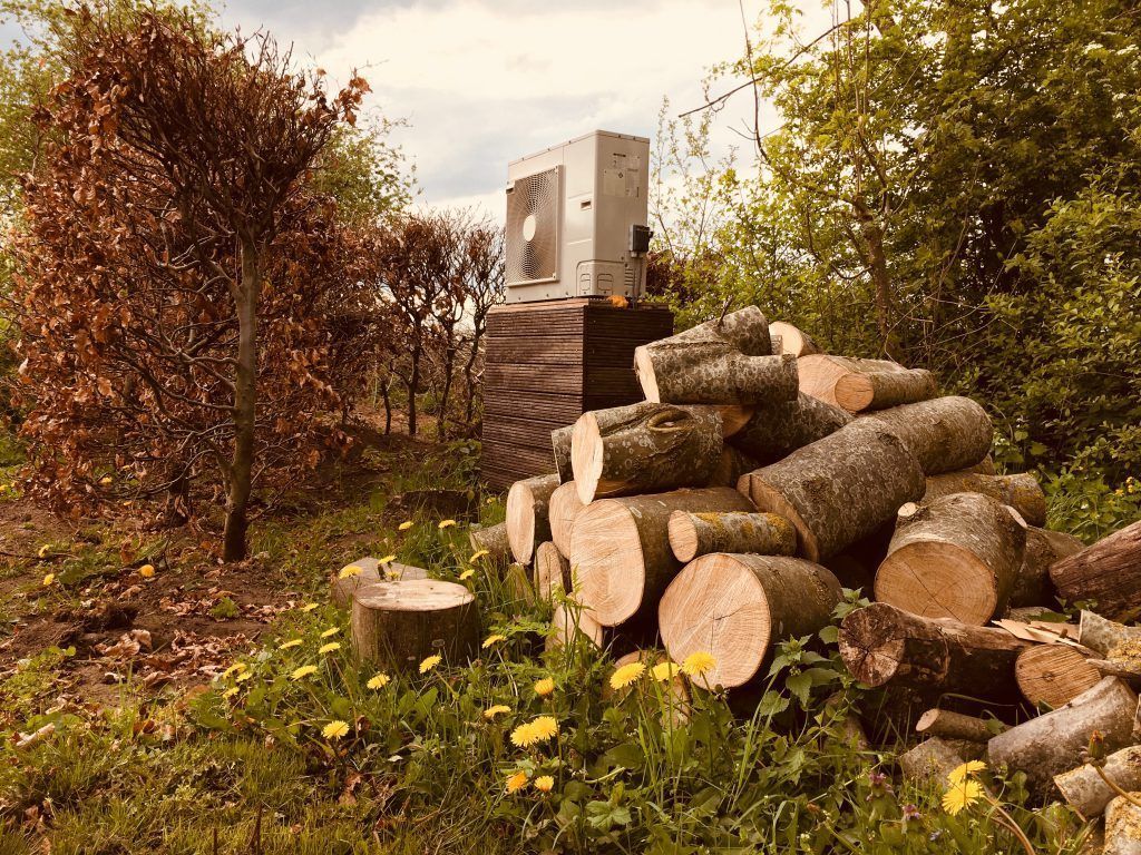 A stack of logs with a large air conditioning unit on top, surrounded by trees and greenery.