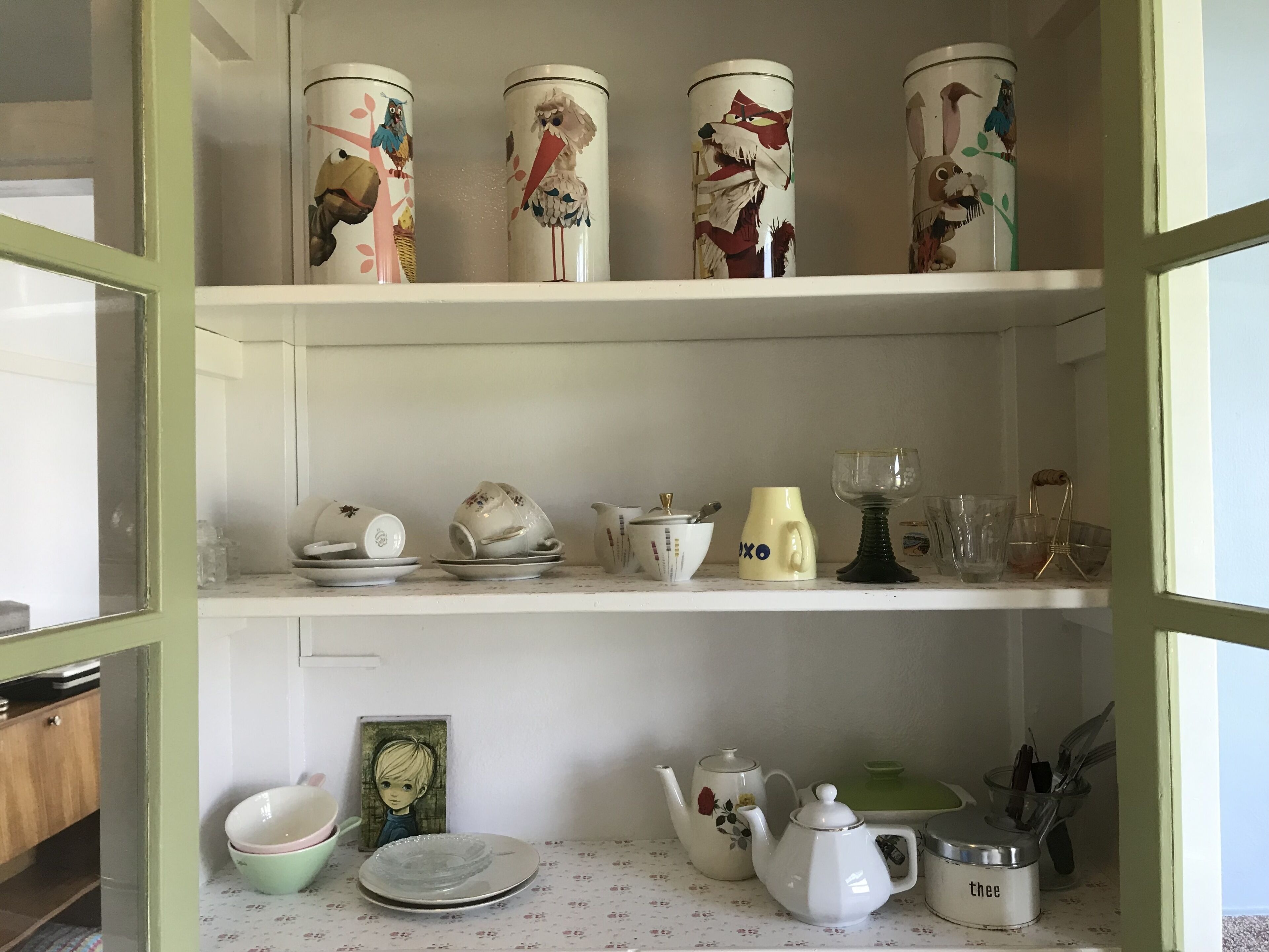 Een keuken met witte kastjes, een tafelkleed met bloemenprint en divers keukengerei, waaronder theepotten, kopjes en een karaf.