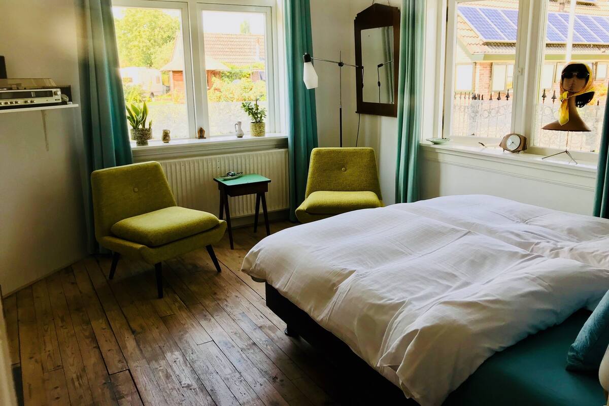 Een slaapkamer met een bed, twee stoelen, een klein tafeltje, een radiator en een raam met gordijnen.