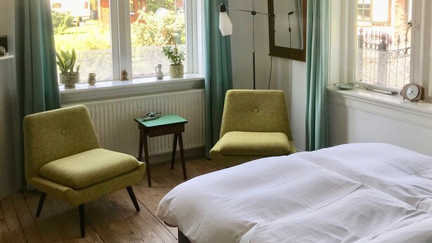 Ein Schlafzimmer mit einem Bett, zwei Stühlen, einem kleinen Tisch, einer Pendelleuchte und einem Fenster mit Vorhängen.
