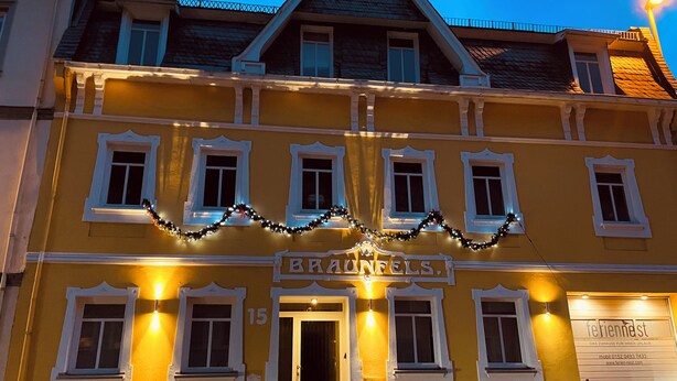 Un edificio giallo con un cartello che riporta la scritta "BRANFELS". Presenta diverse finestre e una porta, con luci sulla facciata.