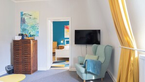 Una stanza con un ampio settimino in legno, una poltrona color petrolio, un televisore a schermo piatto e un quadro colorato appeso al muro.