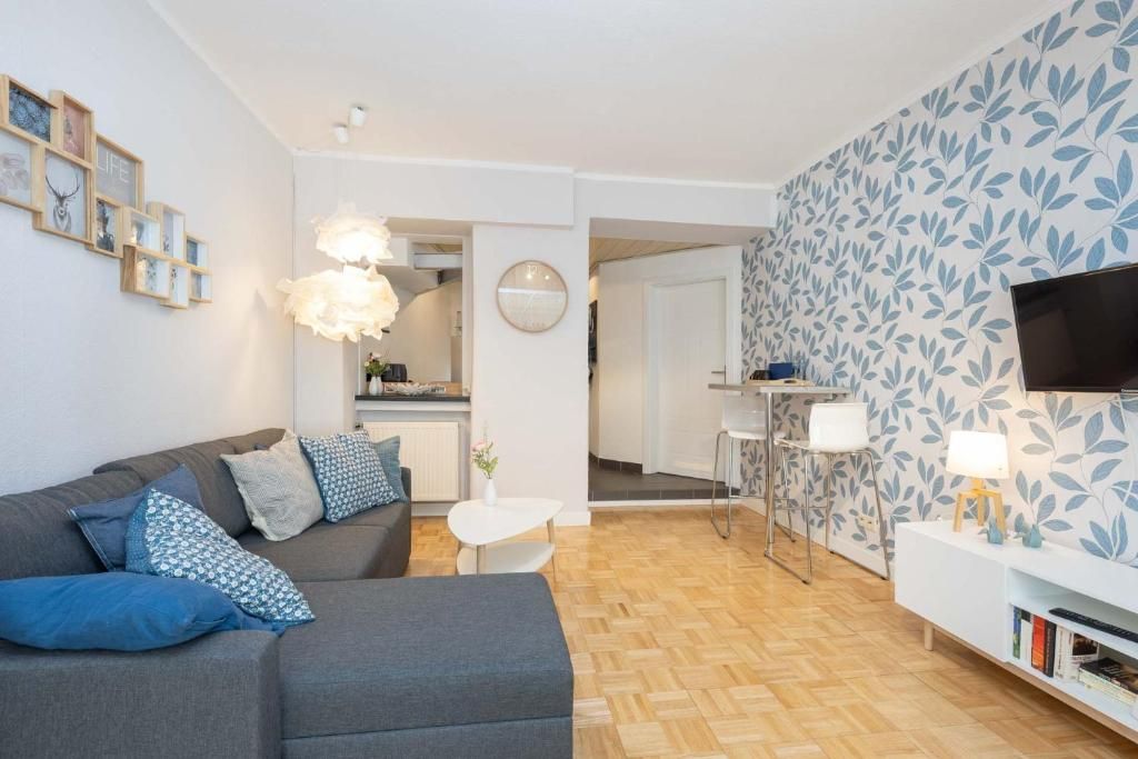 Un salon moderne avec un canapé gris, une petite table blanche, un téléviseur fixé au mur et un papier peint à motifs.