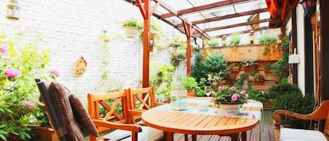 A wooden deck with a round table and chairs, surrounded by potted plants and a brick wall.