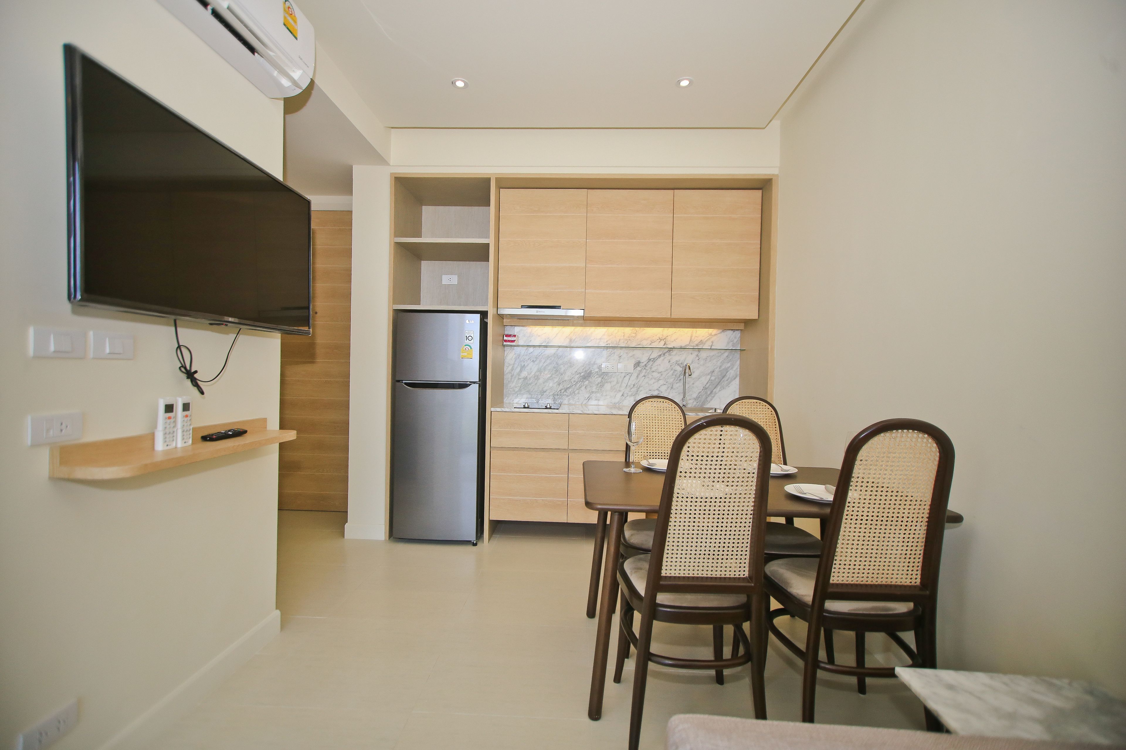 Cocina compacta con refrigerador empotrado, microondas y televisor de pantalla plana montado en la pared. Cuenta con una mesa de comedor con cuatro sillas y un estante pequeño con un teléfono y un control remoto.