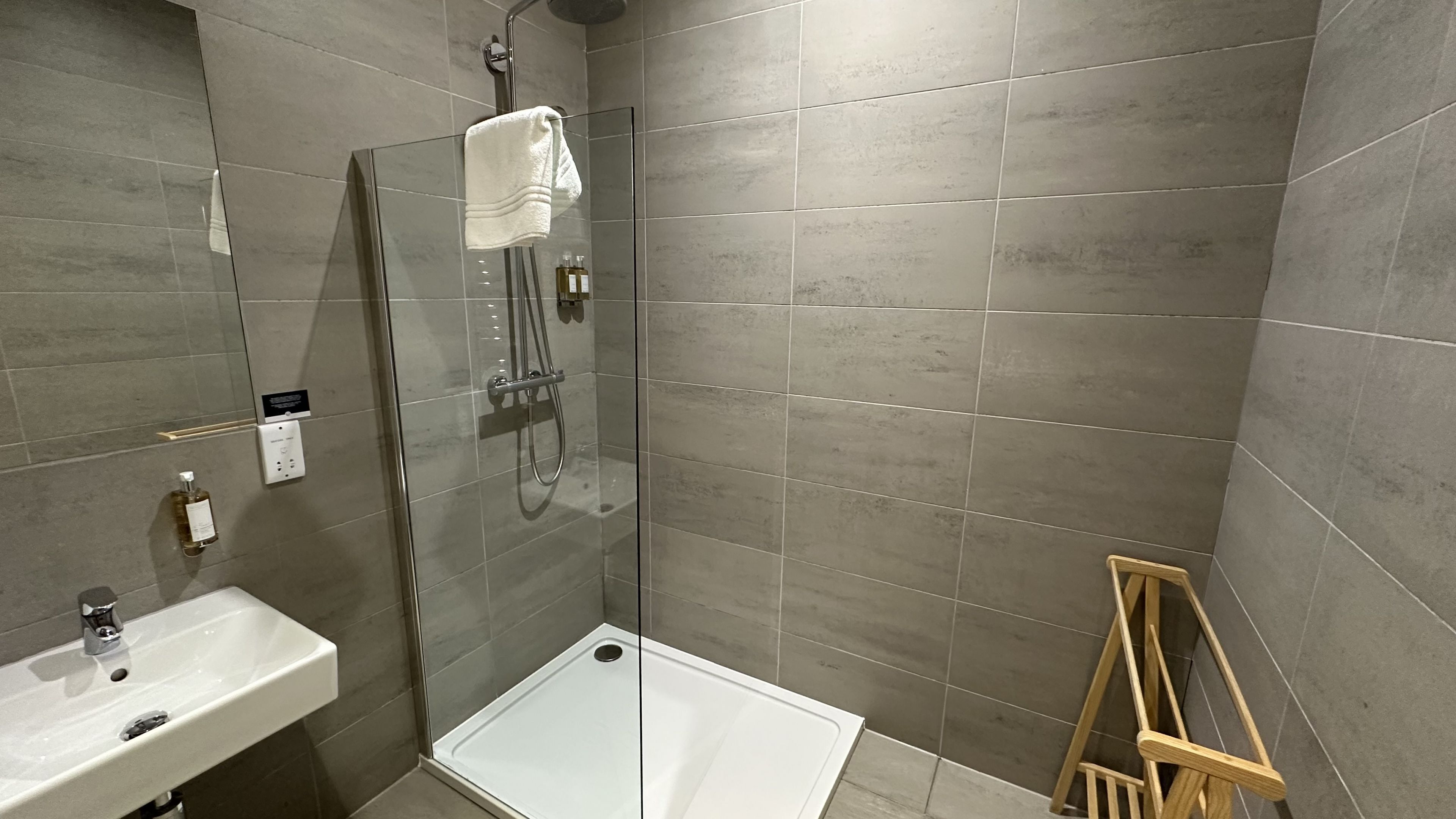 A modern bathroom with a glass shower enclosure, a white sink, and a wooden stool.