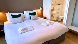 A neatly made bed with white linens and green pillows, a bedside table with a lamp, and a view of a bathroom with a sink and toilet.