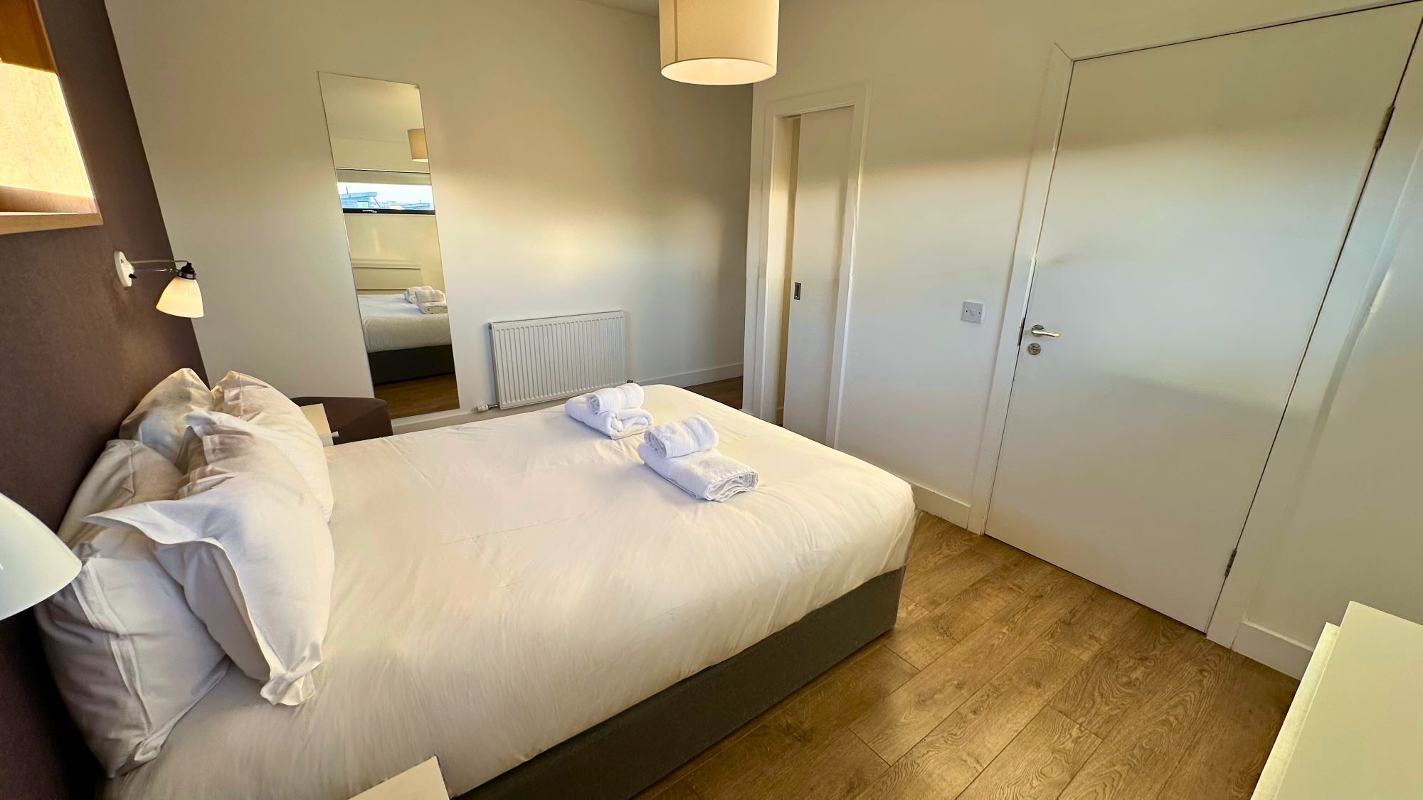Une chambre d’hôtel avec un lit, une lampe de chevet, un miroir, un radiateur et une porte.