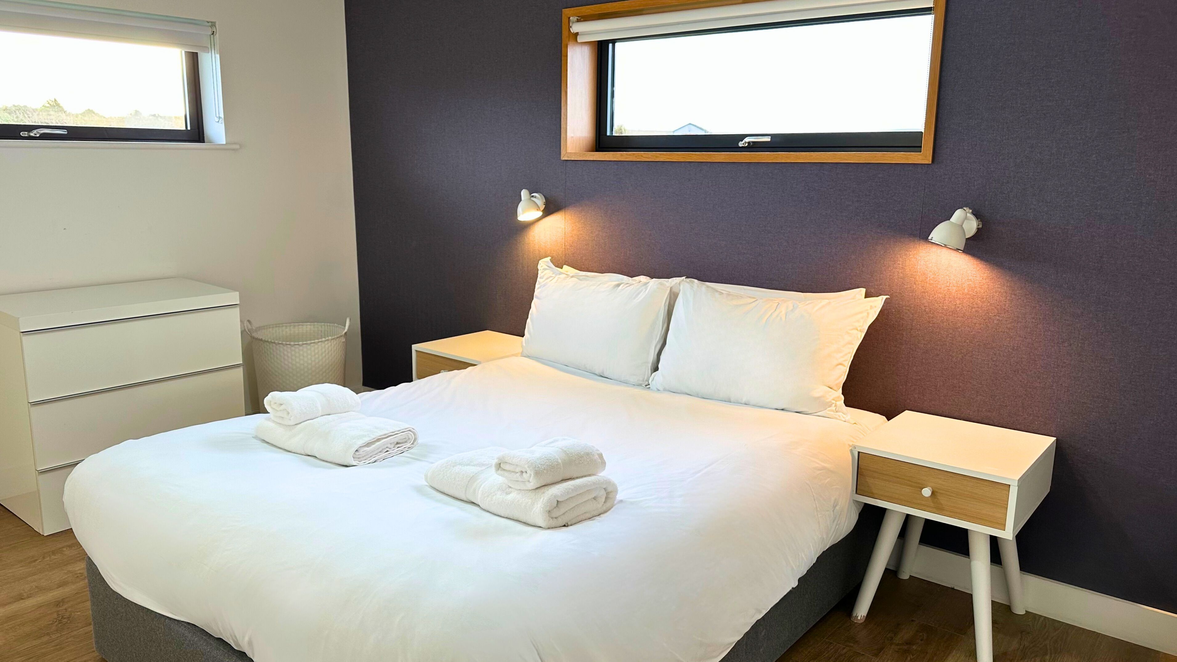 A neatly made bed with white linens and towels, a bedside table with a lamp, a nightstand, and a window with a view of the outdoors.