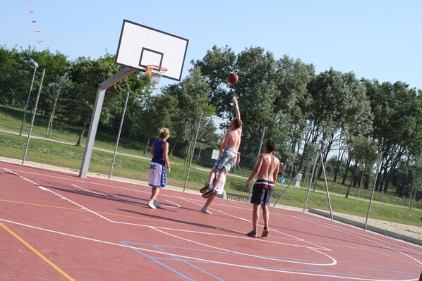 Una cancha de baloncesto al aire libre con un aro, una cerca de malla y árboles de fondo.