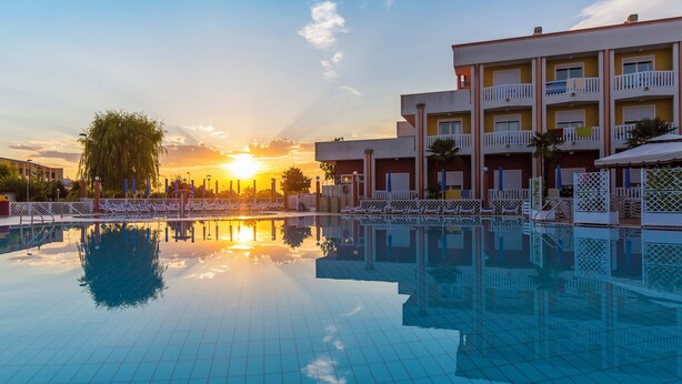 A swimming pool with a sunset reflection, a building with balconies, and a clear sky. - Hotel Olympus (Caorle)