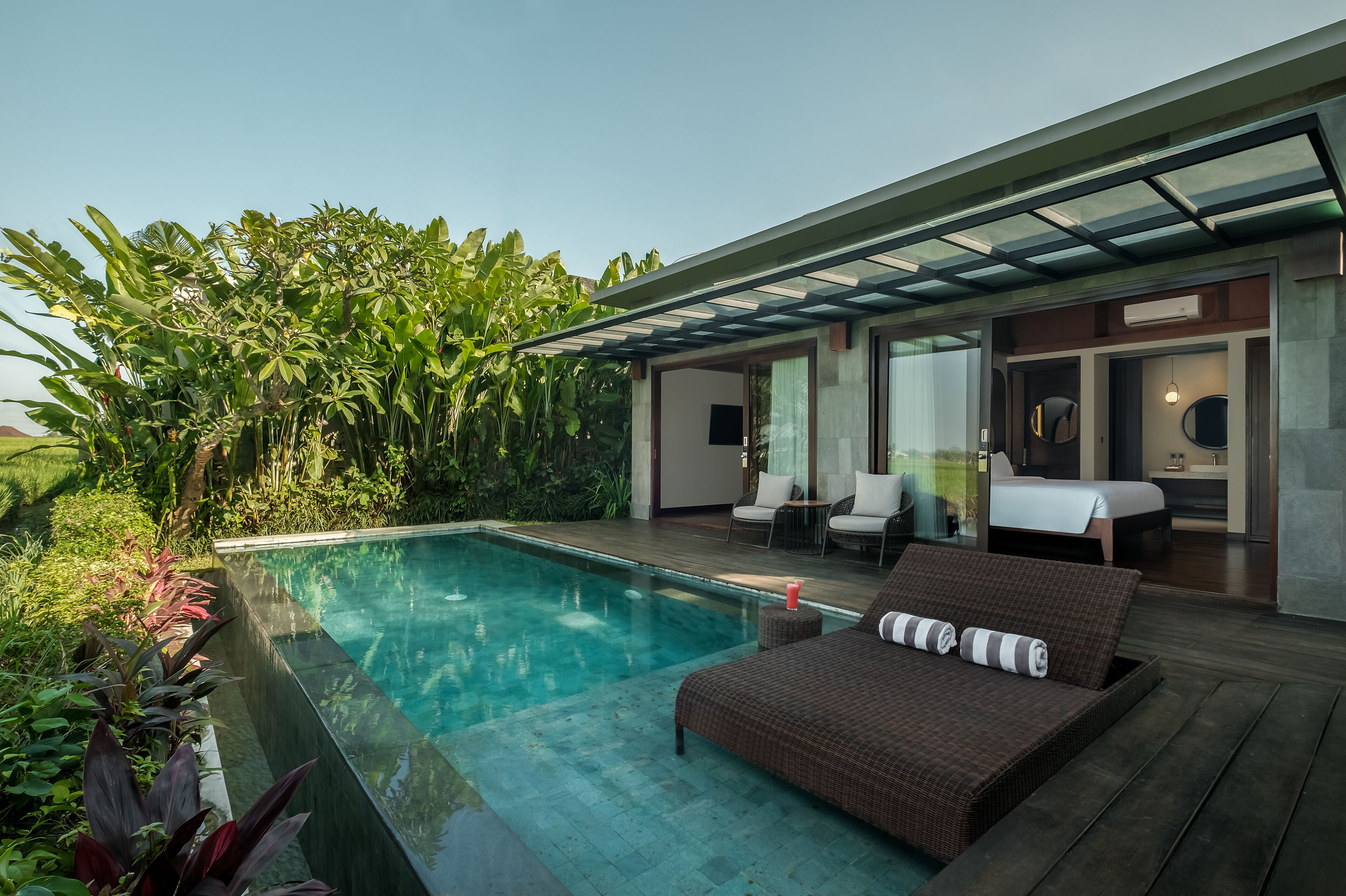 A modern outdoor pool area with a lounge chair and a bed, surrounded by lush greenery.