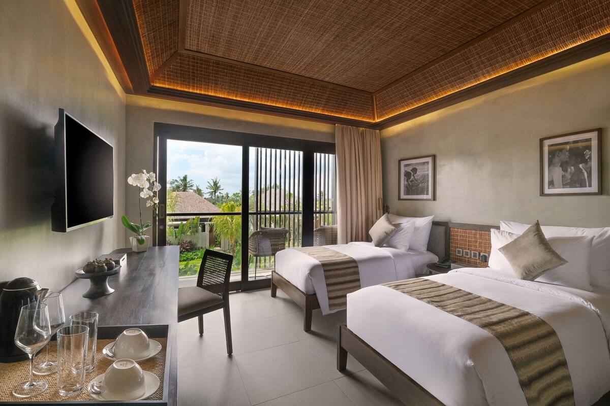 A hotel room with two beds, a desk with a chair, a TV, and a view of the outdoors.