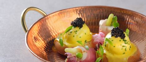 A copper bowl with a handle containing eggs benedict garnished with caviar, green onions, and herbs.