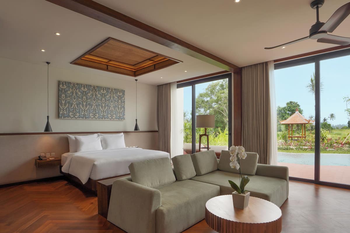 A modern hotel room with a large bed, a green sofa, a round wooden coffee table, and a view of a garden and pool.