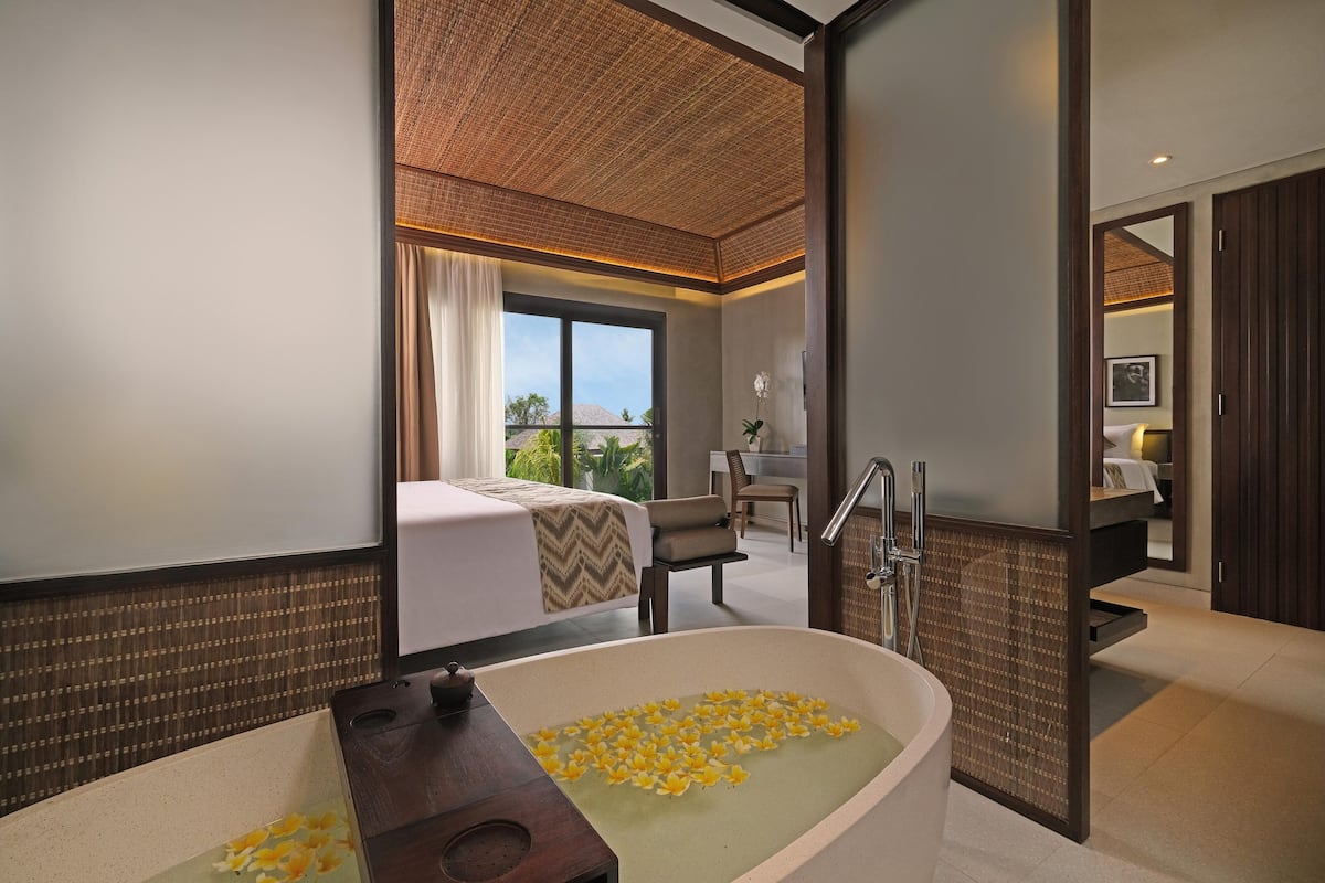A modern hotel room with a large bathtub filled with yellow flowers, a bed with a beige and brown patterned bedspread, and a view of the outdoors through a large window.