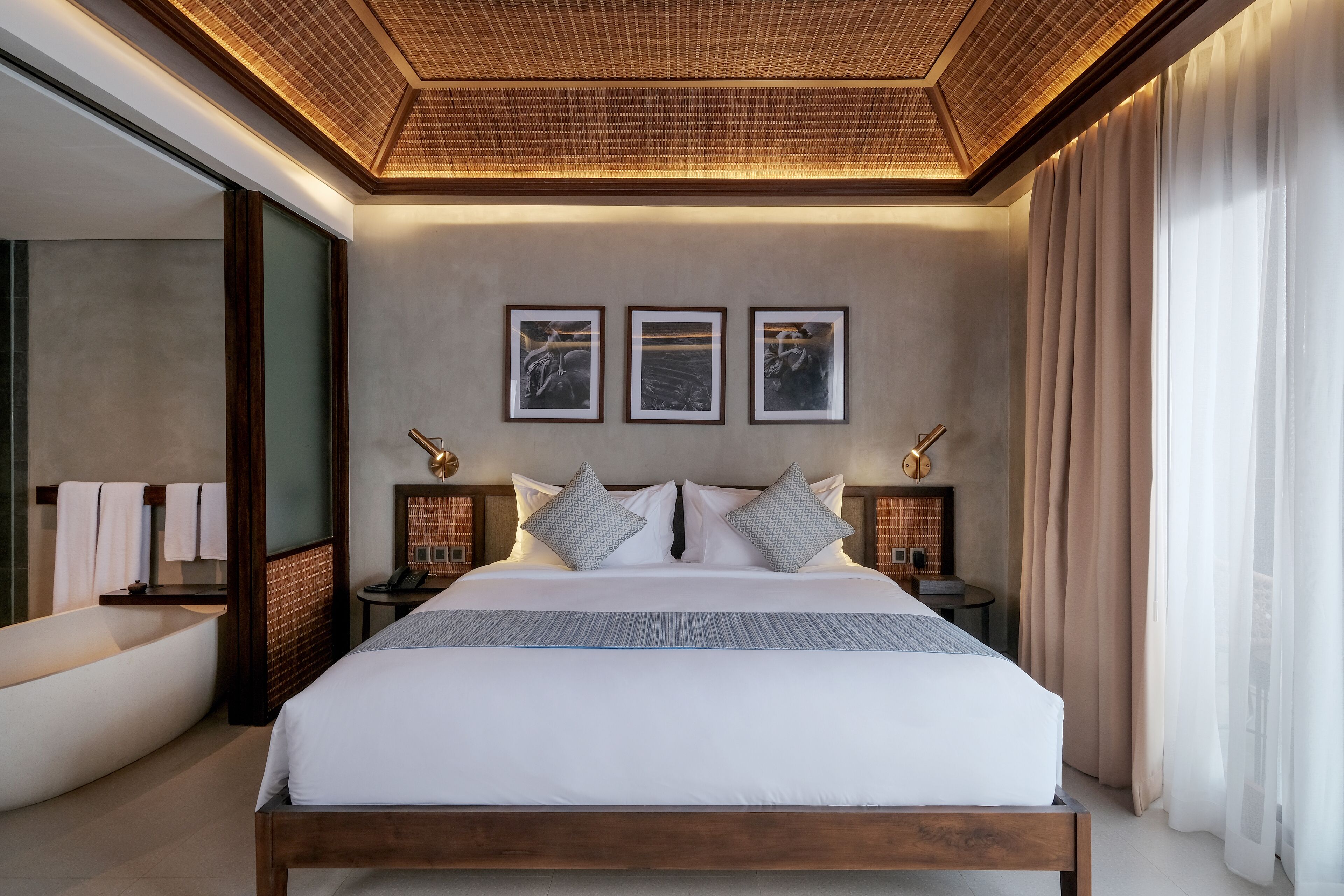 A modern hotel room with a large bed, a wooden headboard, a built-in bathtub, and framed artwork on the wall.