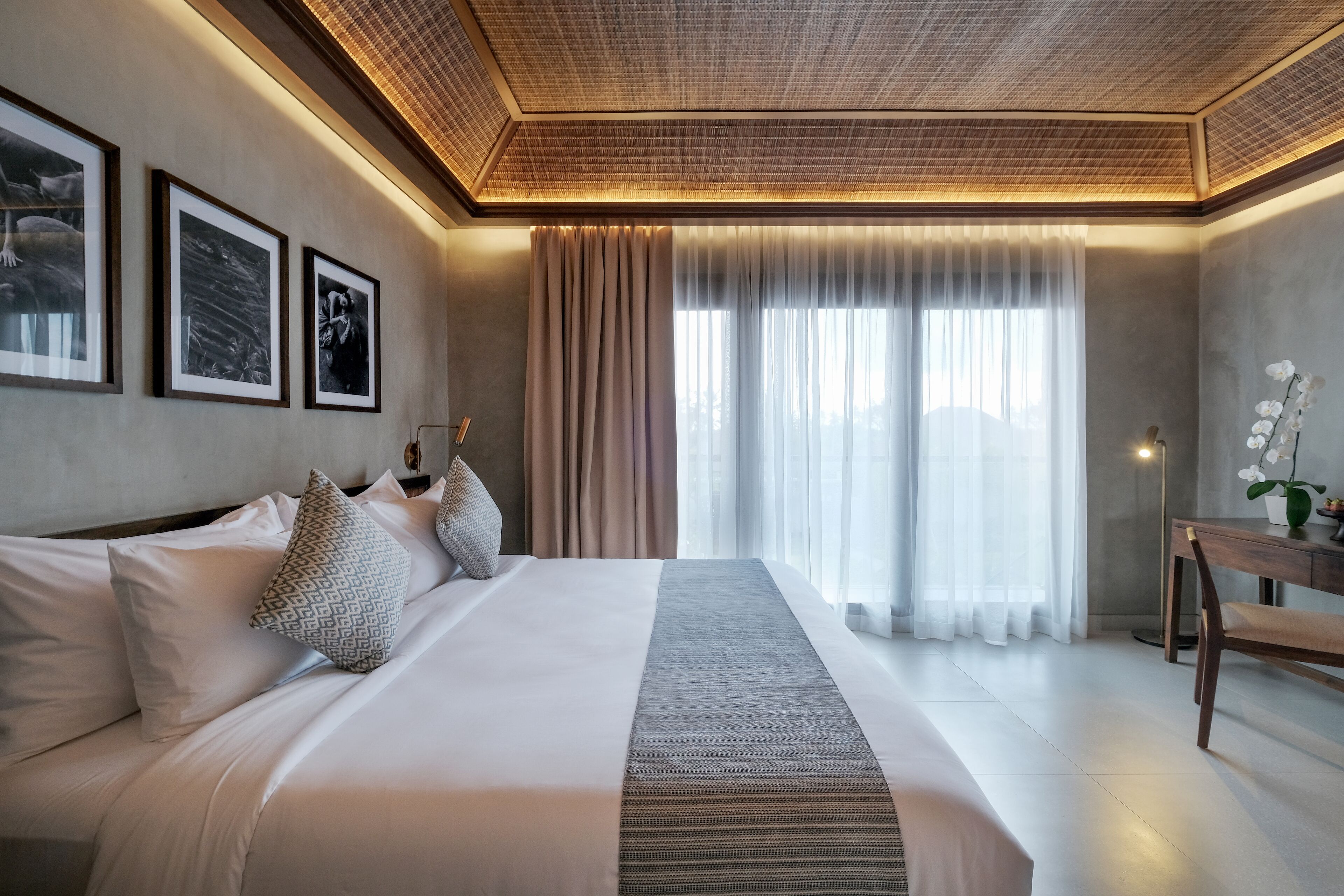 A modern hotel room with a large bed, a desk, and a view of the city through sheer curtains.