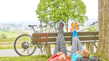 Two people lying on a bench with bicycles parked behind them, in a grassy area with a tree and a view of a distant landscape.