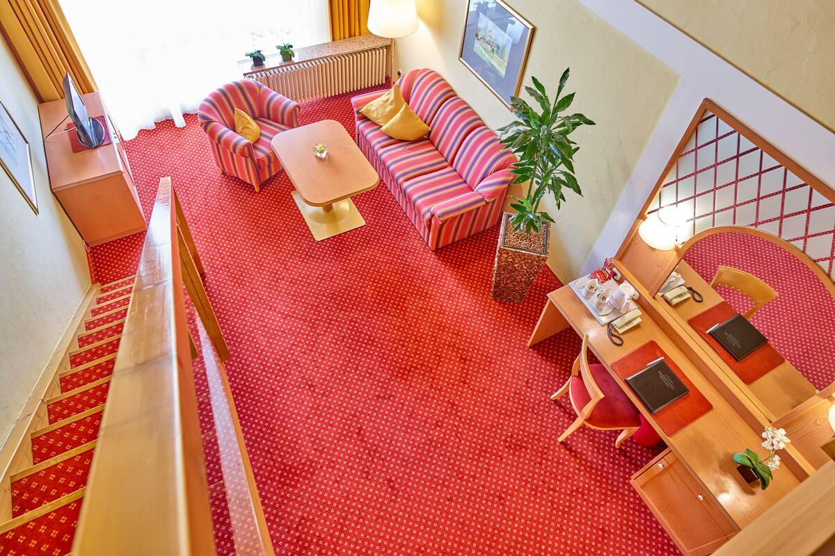 A hotel room with a red carpet, striped sofas, a small table, a potted plant, a desk with a lamp, and a framed picture on the wall.