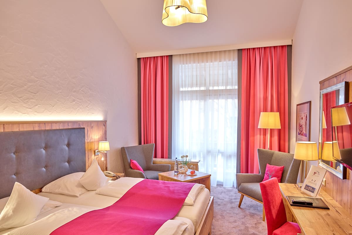 A hotel room with a large bed, two armchairs, a wooden desk, and a large window with curtains.