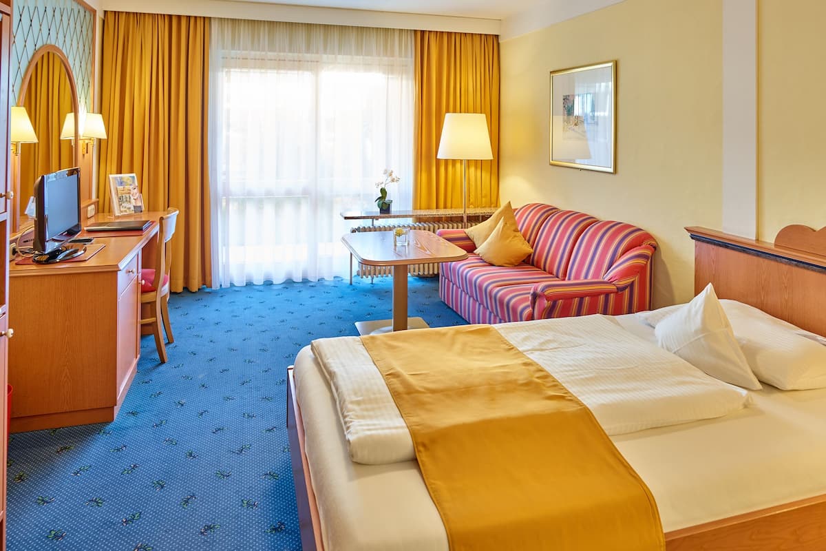A hotel room with a bed, a desk with a computer, a chair, a sofa, and a small table with a lamp.