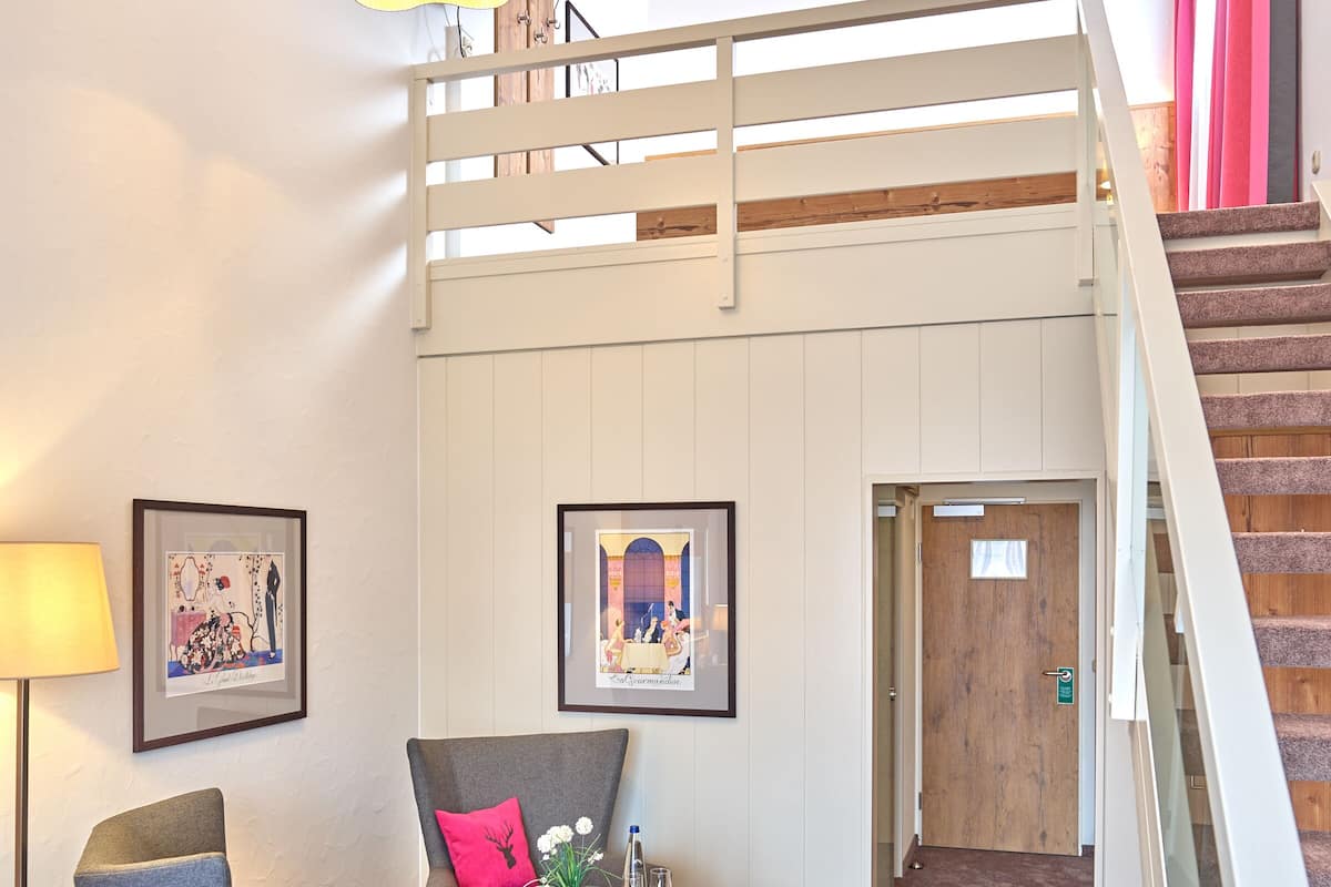A modern interior with a loft-style bed, a wooden coffee table with a vase and fruit, a grey armchair, and a staircase with a wooden railing.