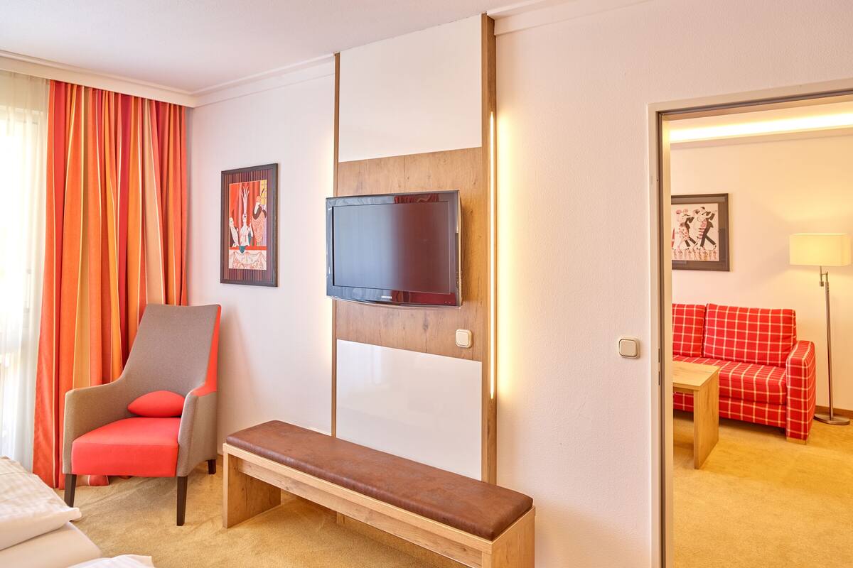 A hotel room with a bed, a red armchair, a wooden bench, a television mounted on a wooden panel, and a red checkered sofa in the adjacent room.