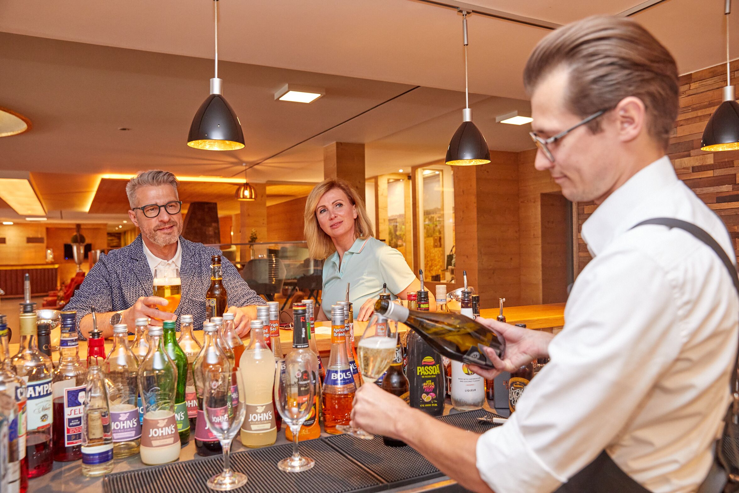 Un espace bar avec de nombreuses bouteilles et des verres, un barman qui sert des boissons et un décor intérieur en bois.