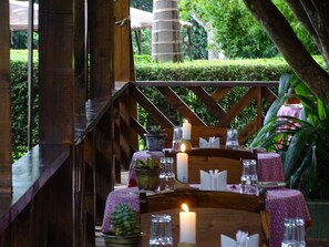 Un área de comedor exterior de madera con mesas dispuestas para una comida, rodeada de follaje y una estructura de madera.